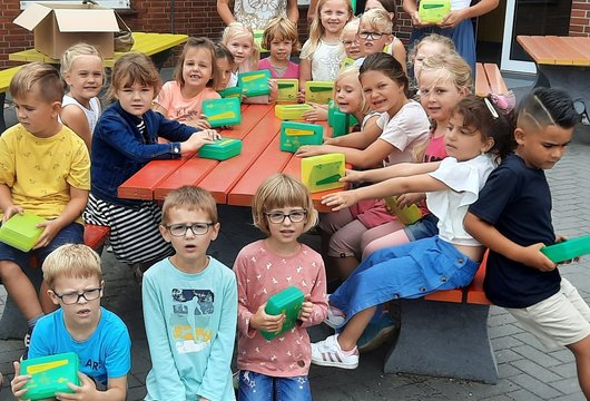 Kinder sitzen um einen Tisch mit grünen und gelben Plastik-Brotdosen.