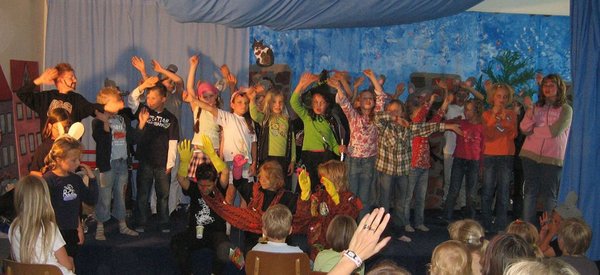 Eine Gruppe von Kindern winkt dem Publikum von einer Bühne aus zu.